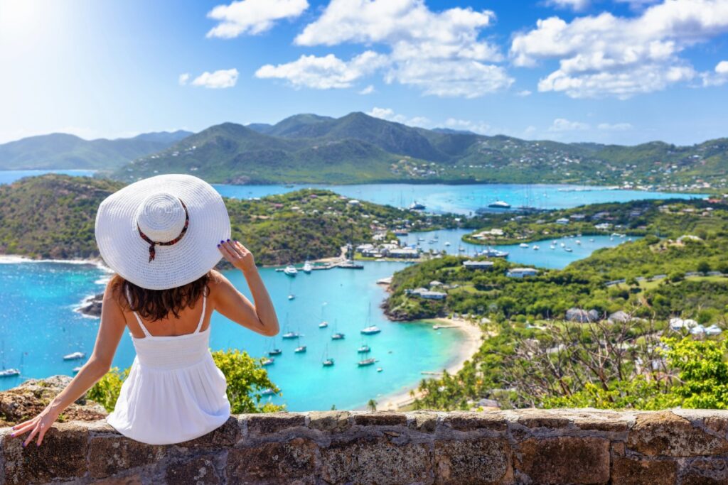 A tourist woman in a white dress enjoys the panoramic view over the landscape of Antigua island with English Harbour and Shirley heights